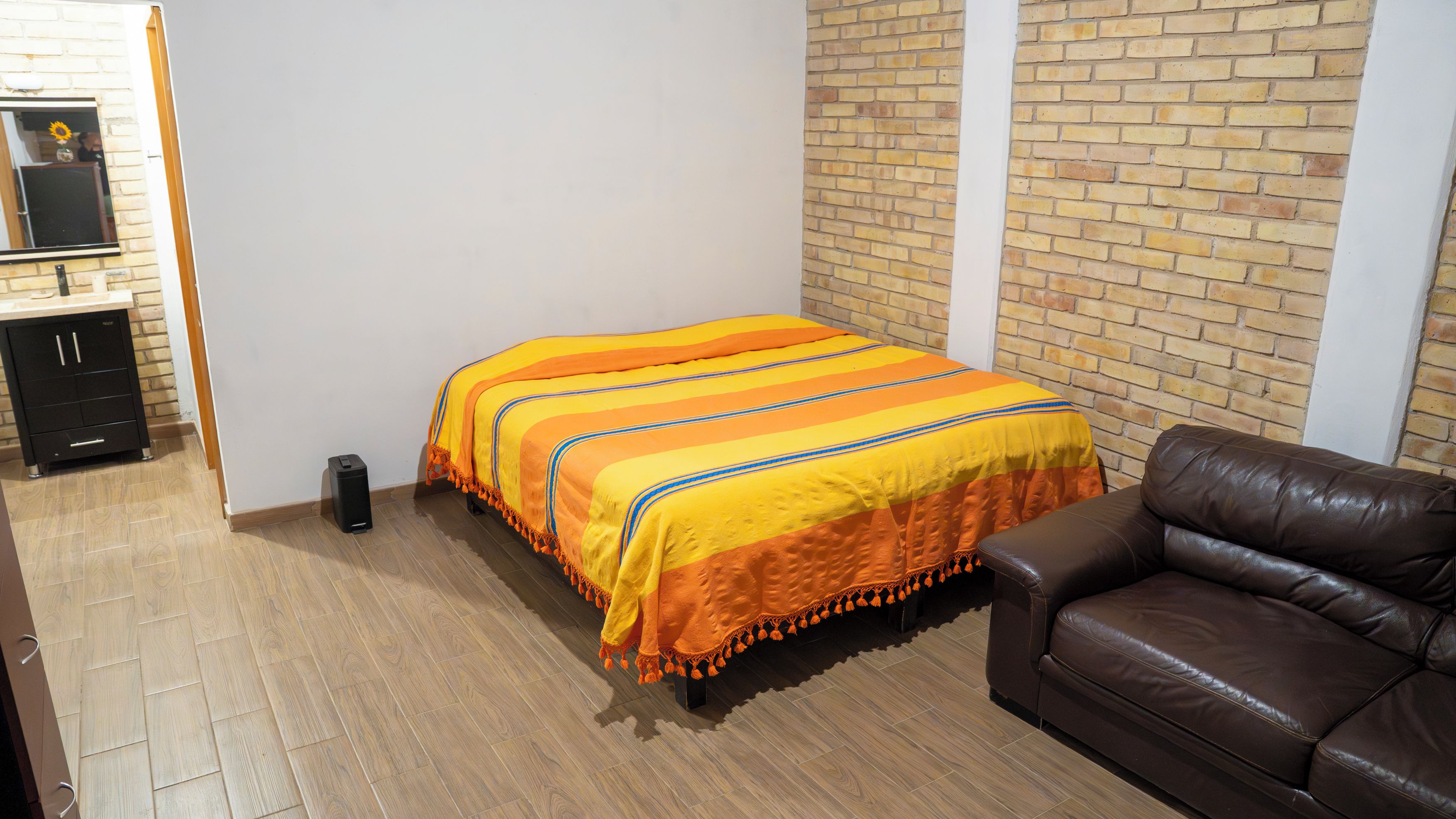 A bedroom with a bed, a brown leather sofa, a small black speaker, and a brick wall.
