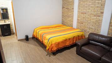 A bedroom with a bed, a brown leather sofa, a small black speaker, and a brick wall.