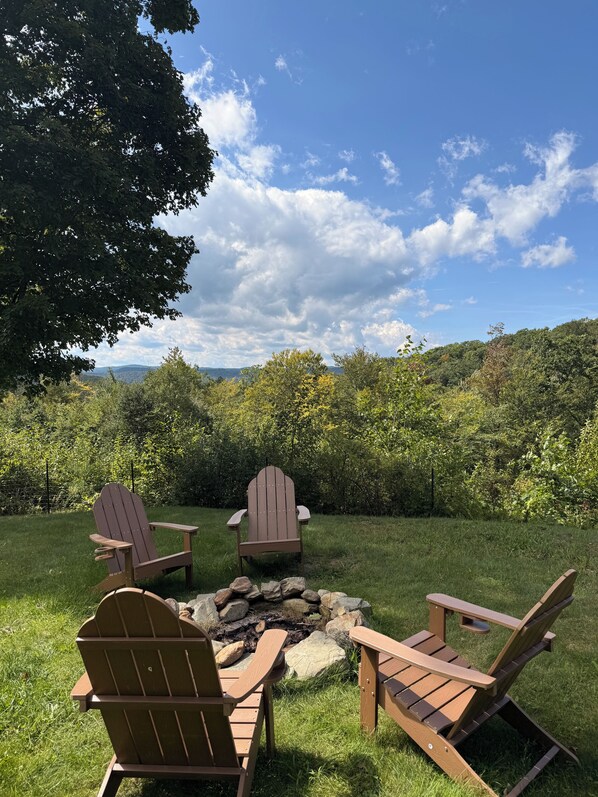 A fire pit with three Adirondack chairs in a grassy outdoor area with a view of a forest and mountains.