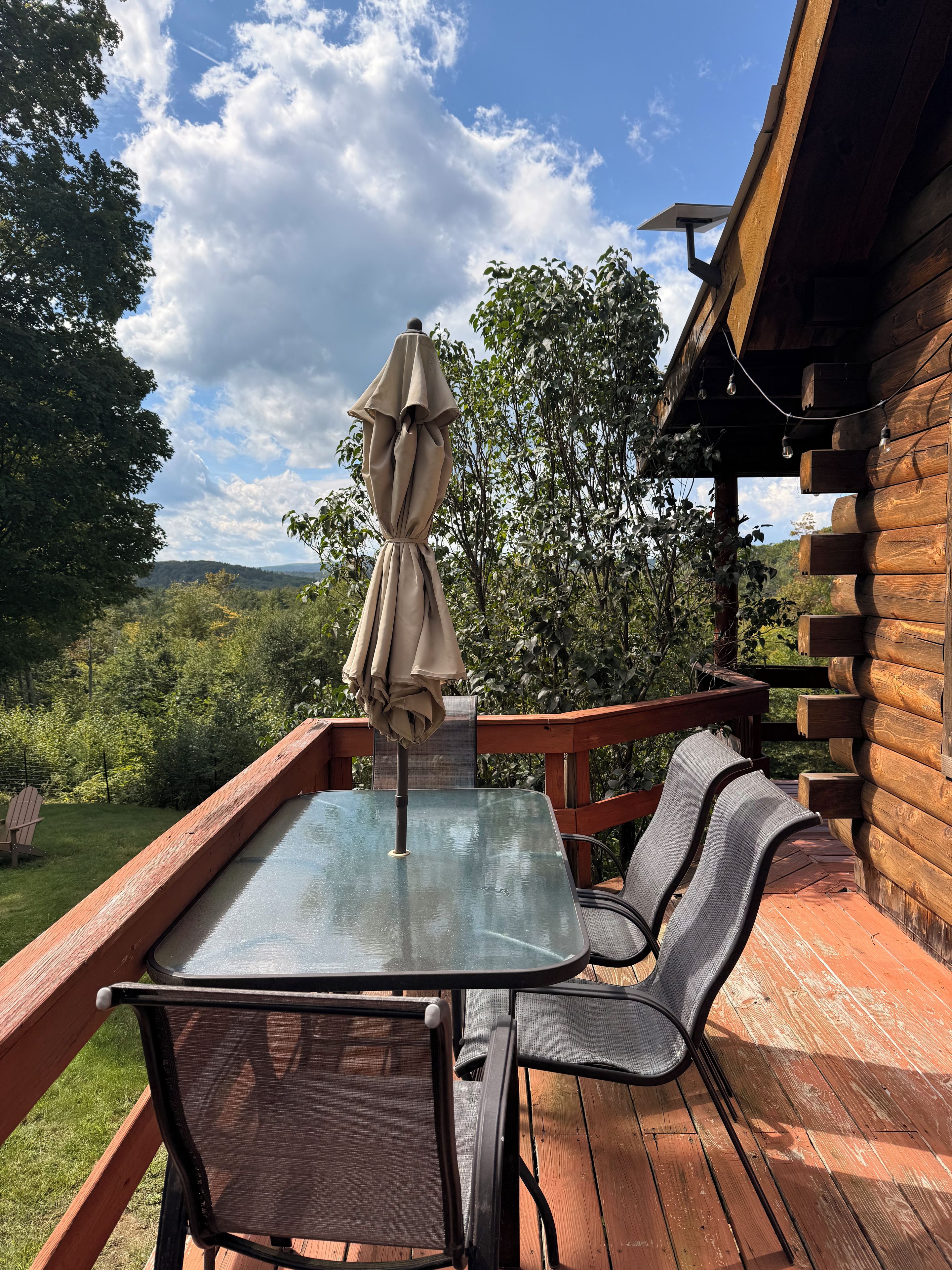 A wooden deck with a glass table and chairs, an umbrella, and a view of the surrounding greenery.