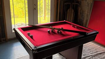 A pool table with a red felt surface, a door with a view of greenery outside, and a wall with a sign indicating a hiking trail.