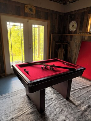 A pool table with a red felt surface, a door with a view of greenery outside, and a wall with a sign indicating a hiking trail.