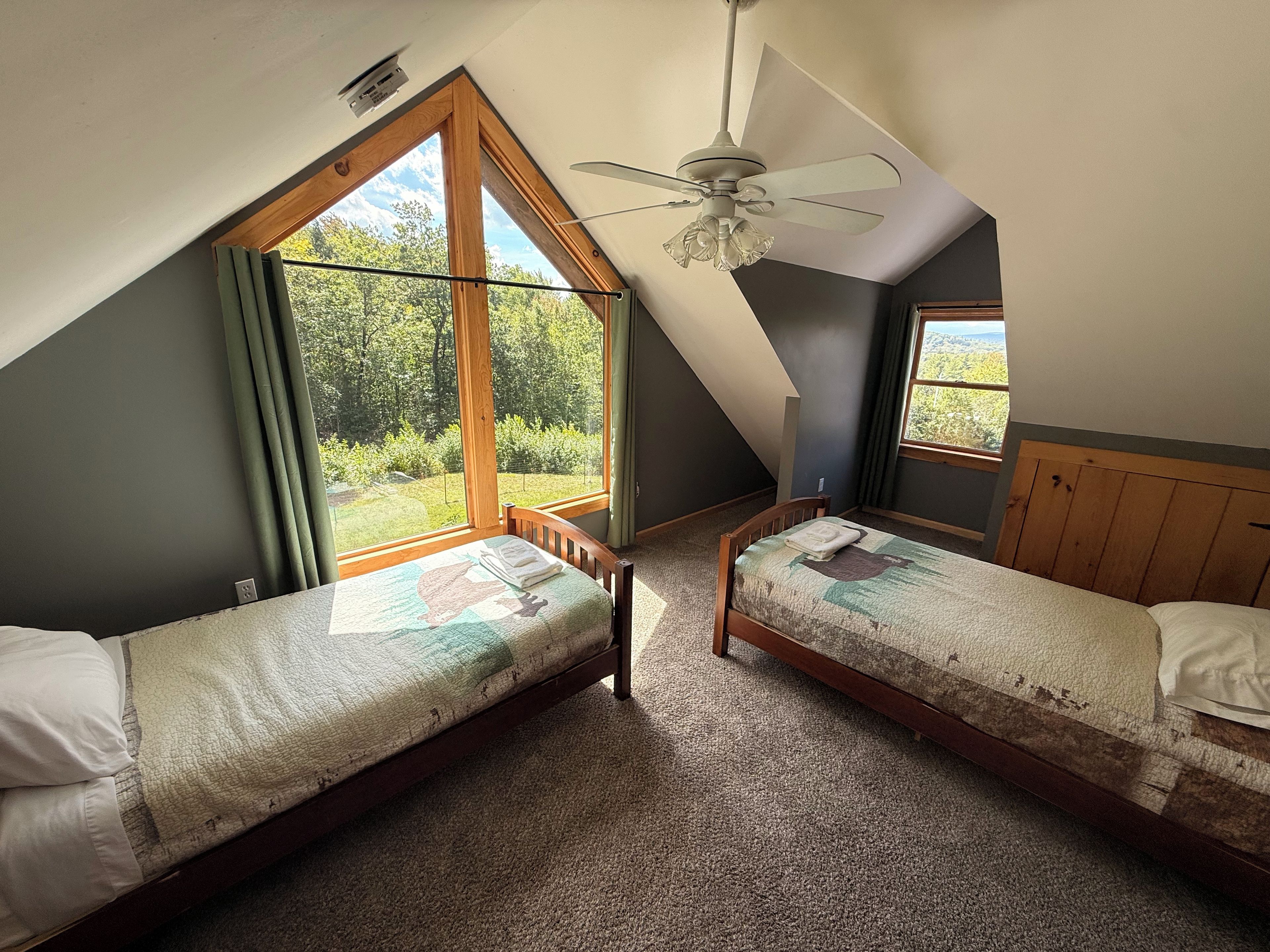Quarto com duas camas de solteiro, ventilador de teto e amplas janelas com vista para o verde.