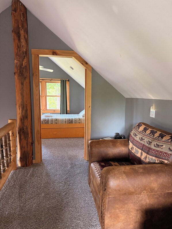 A loft bedroom with a sloped ceiling, a wooden ceiling beam, a bed with a patterned bedspread, and a brown leather sofa with a decorative throw.