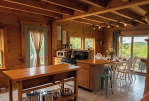 Cocina de cabaña de madera con comedor, vigas de madera, estufa, microondas y vista al exterior.