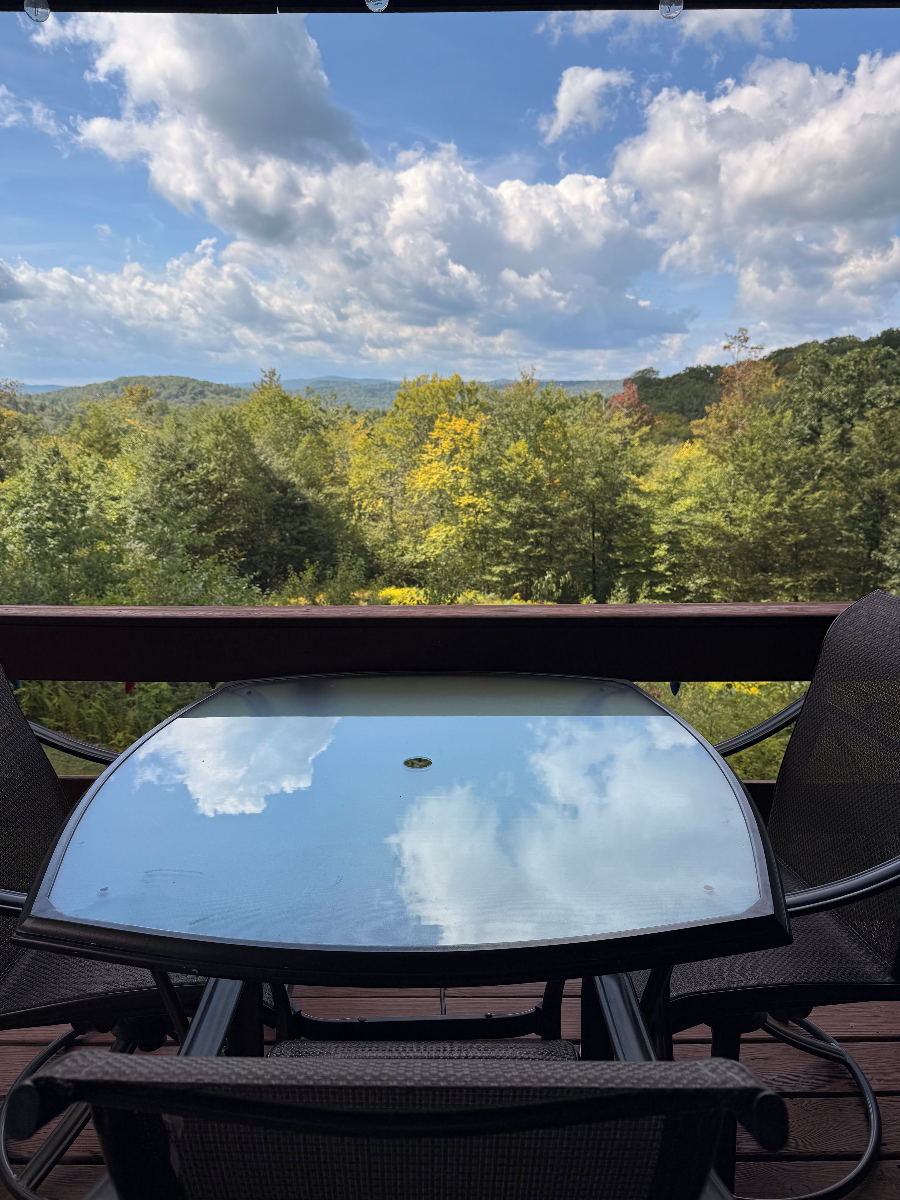 A table with chairs on an outdoor deck, overlooking a forested area with hills in the distance.