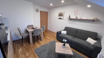 Een moderne woonkamer met een grijze bank, een klein bijzettafeltje en een eethoek met een houten tafel en stoelen.