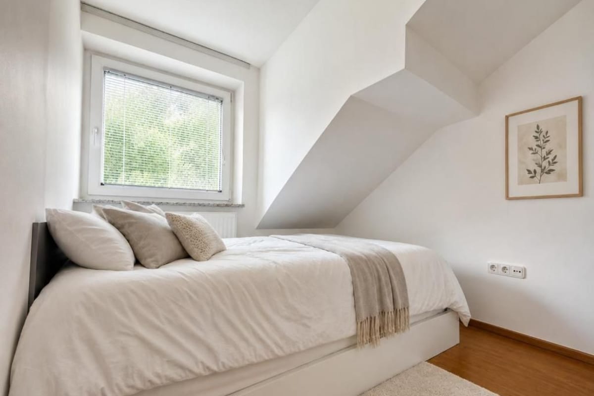 Een slaapkamer met een bed, een raam, een wandcontactdoos en een lijst met een afbeelding.