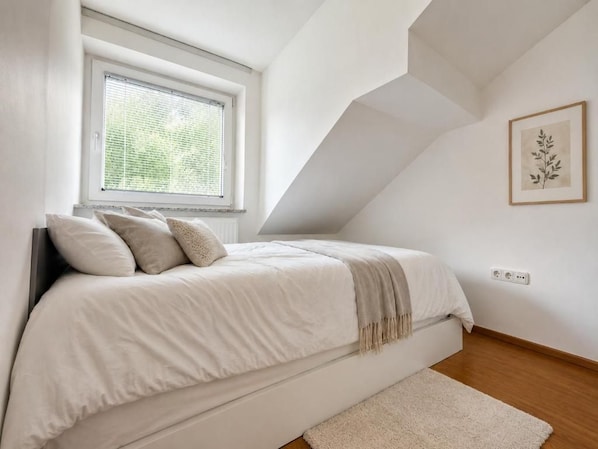 A bedroom with a bed, a window, a wall-mounted power outlet, and a framed picture.