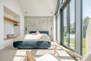 A modern bedroom with a large bed, a wooden bench, a nightstand, and a view of greenery outside. - Ferienhaus für Familien in Fuhlendorf am Hafen (Fuhlendorf)