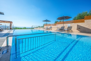 Ein klarer, blauer Swimmingpool mit Liegestühlen und Sonnenschirmen, umgeben von einem Holzzaun und Blick aufs Meer.