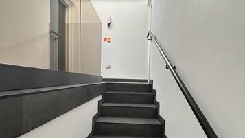 A modern staircase with a glass ceiling, metal handrails, and a white wall with a fire alarm.