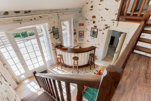 A high-ceilinged room with a wooden bar and stools, a large window, and a staircase. - La Prairie Authenticité et Confort -piscine privée (Villeneuve-les-Bordes)