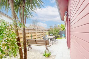Une terrasse avec une table et des chaises en bois, un banc et un petit jardin agrémenté d’un palmier et de plantes à fleurs.