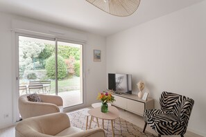 A modern living room with a large sliding glass door leading to a garden, a flat-screen TV on a white cabinet, a zebra-patterned chair, and a small round table with a vase of flowers. - Pic épeiche - proche de la plage (Pléneuf-Val-André)