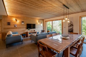A modern interior with a wooden ceiling, a dining area with a large table and chairs, a sofa, and a television mounted on the wall. - Chalet Soline La Glière - Appartement avec jardin (Samoëns)