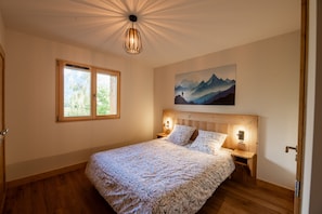 A bedroom with a wooden headboard, a bed with patterned bedding, a nightstand, a wall-mounted lamp, and a window with a view of greenery. - Chalet Soline La Glière - Appartement avec jardin (Samoëns)