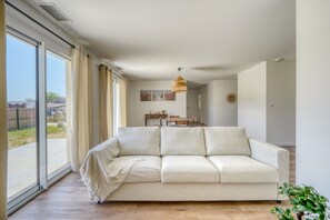A spacious living room with a large sofa, a dining area with a table and chairs, and a sliding glass door leading to an outdoor area. - La Casa Romarin - Proche Libourne (Galgon)