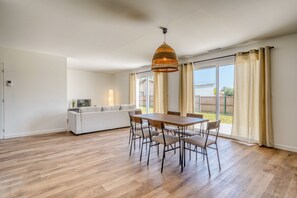 Un salón espacioso con suelo de madera, una mesa de comedor con sillas, un sofá y un amplio ventanal con cortinas.
