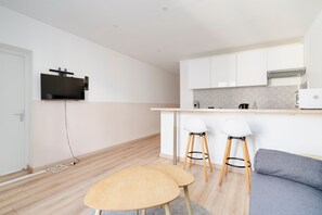 Een moderne keuken met witte kasten, een ingebouwde oven en een flatscreen televisie boven het aanrecht. In de woonkamer staan twee houten krukken en een grijze bank.