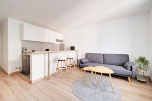 A modern kitchen with white cabinets, a microwave, and a coffee machine. There is a grey sofa and a small round table with a grey rug in the living area. - Le Mozart nancéen - proche du centre-ville (Nancy)