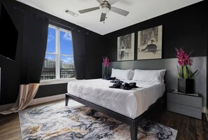 Ein modernes Schlafzimmer mit Bett, Deckenventilator, Wandkunst und Blick auf die Stadt durch ein Fenster.