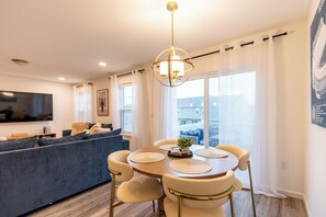 A modern living room with a sofa, dining table, and chairs, a flat-screen TV, and a large window with curtains. - 2 Back-to-Back Homes | Sleeps 18 | Near PSU (Bellefonte)