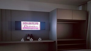 Une chambre à coucher avec un lit, un téléviseur fixé au mur et une étagère où sont posées des bouteilles et des figurines.