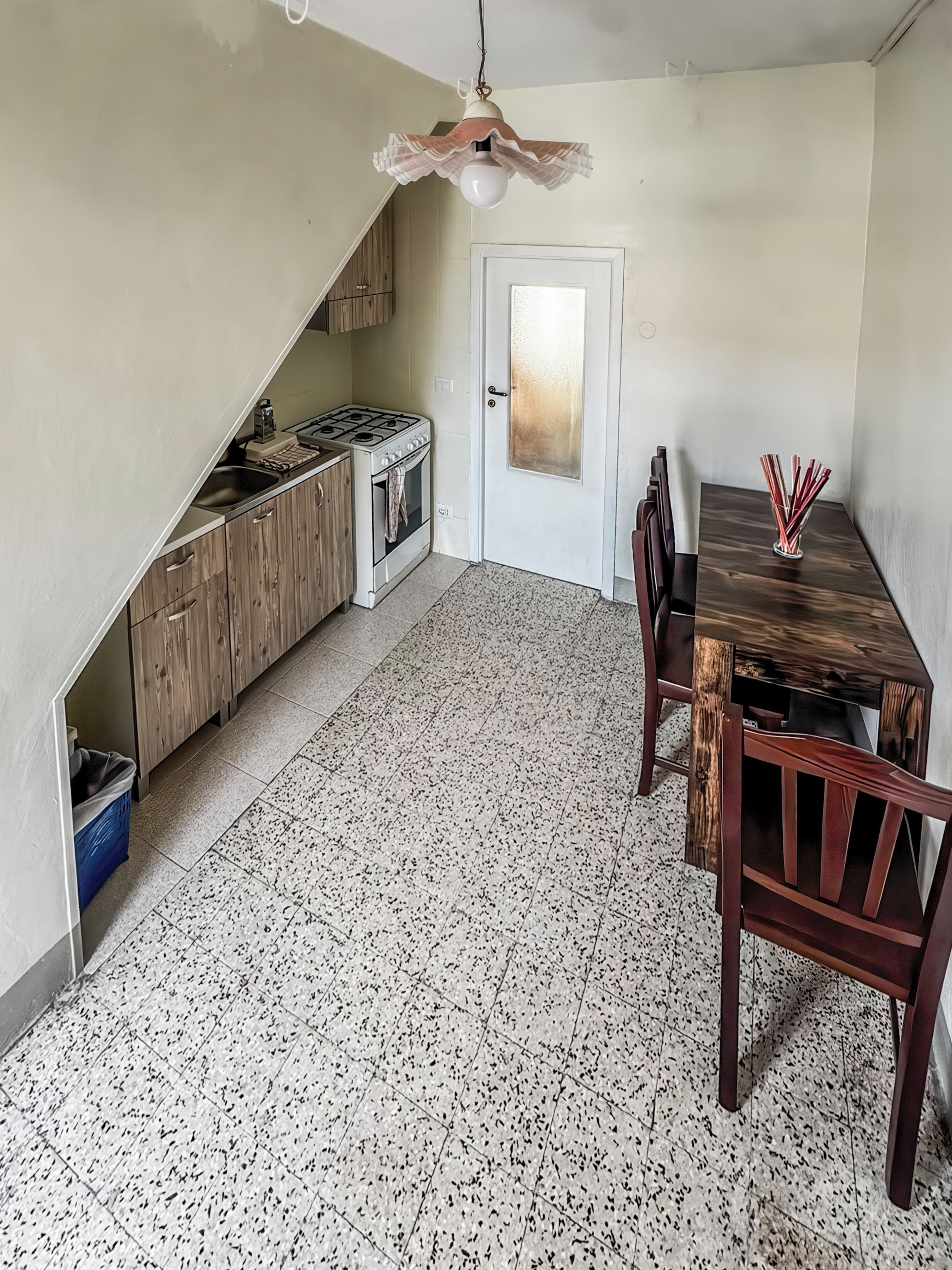 A compact kitchen with a gas stove, wooden cabinets, a dining table with chairs, and a speckled floor.