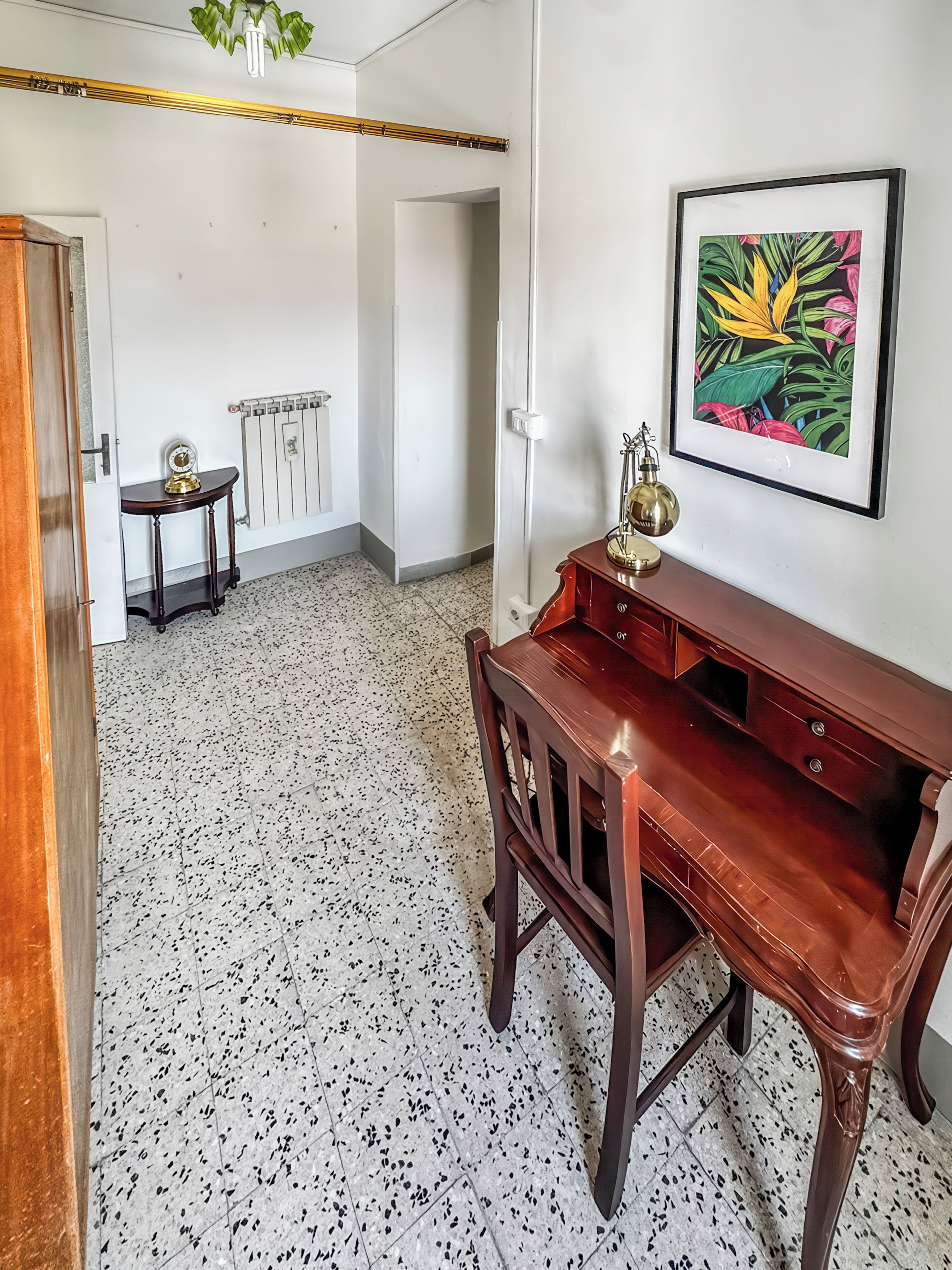 Una habitación con un escritorio de madera, una silla, un radiador, un cuadro en la pared y una puerta que da a otra habitación.