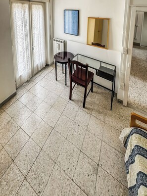 A room with a bed, a table with a chair, a radiator, a mirror, and a window with curtains.