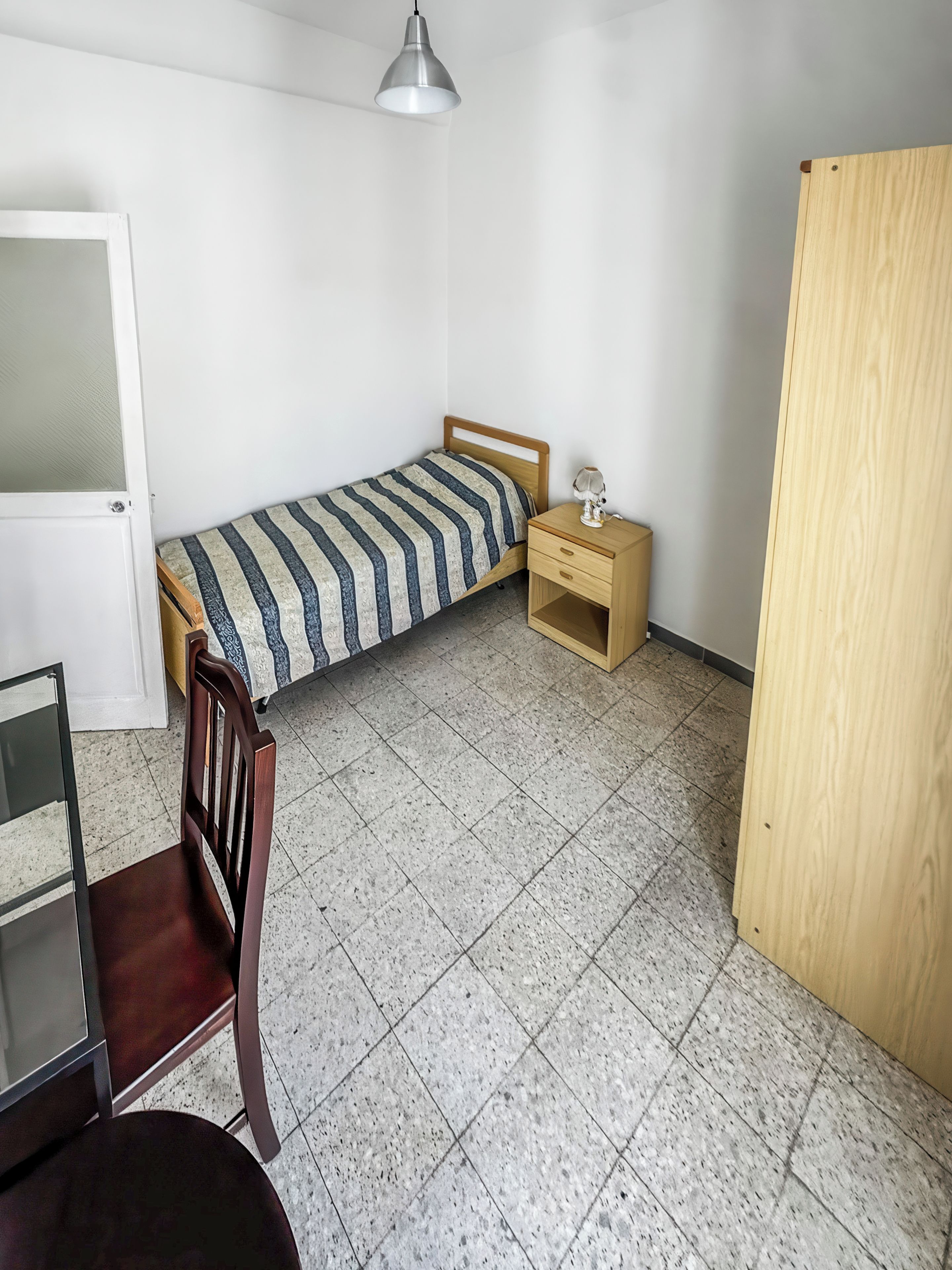 A small, sparsely furnished room with a bed, a chair, a desk, and a wardrobe.