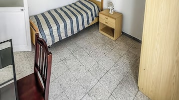A small, sparsely furnished room with a bed, a chair, a desk, and a wardrobe.
