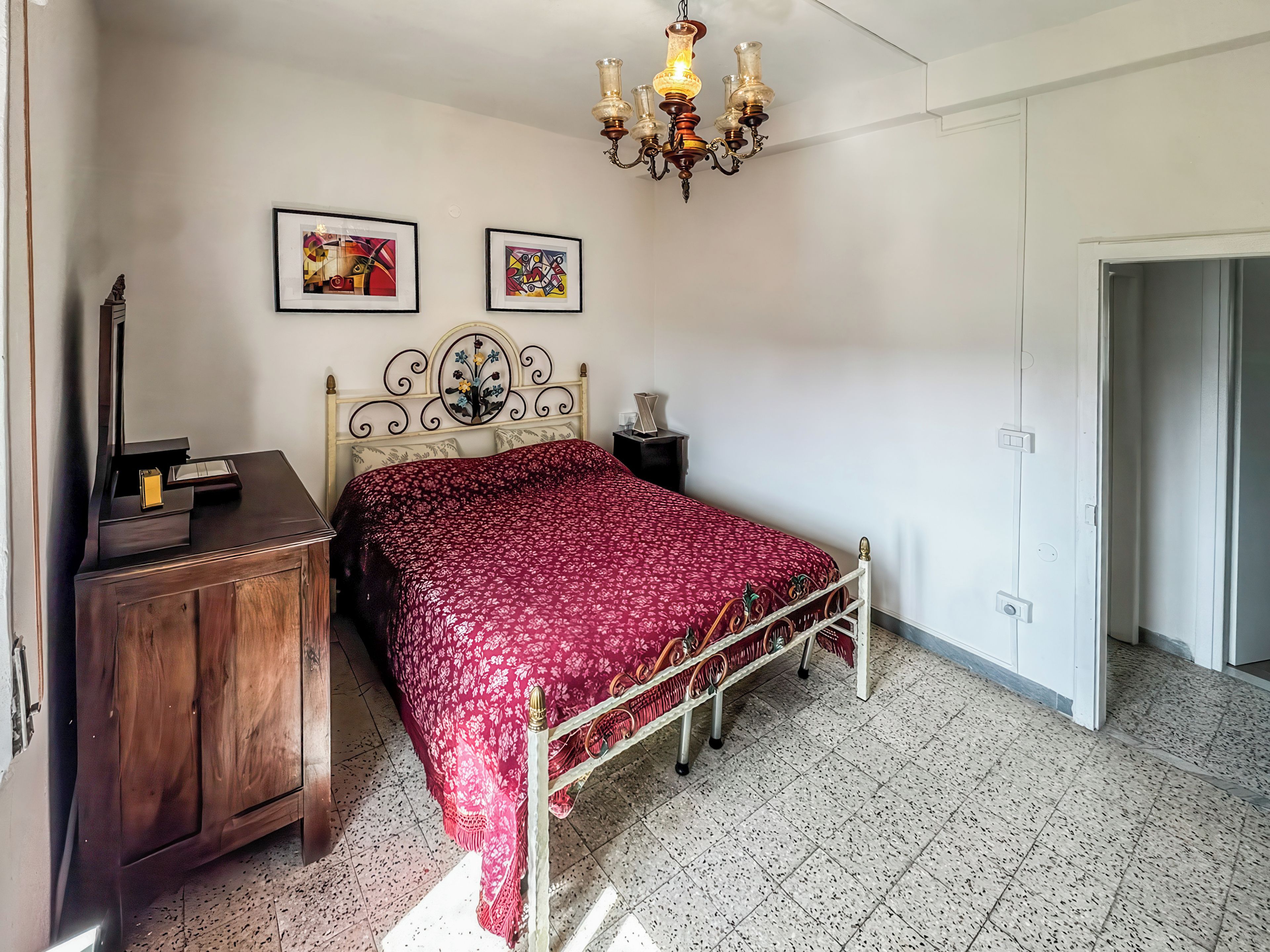 Ein Schlafzimmer mit Bett, Schminktisch, einer Kandelaber und zwei gerahmten Kunstwerken an der Wand.