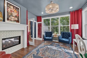 A room with a fireplace, a large window with red curtains, a blue armchair, and a patterned rug. - Pet Friendly-Baileys Harbor Home with Fire Pit (Baileys Harbor)