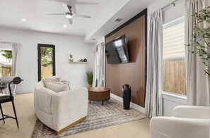 A modern living room with a sofa, a TV mounted on the wall, and a ceiling fan. - “Luxury Living Meets Comfort – 4BR Dream Stay” (Houston)