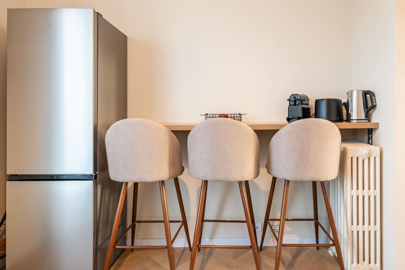 Une petite cuisine équipée d’un réfrigérateur en acier inoxydable, de trois tabourets de bar rembourrés, d’une machine à café et d’une bouilloire, le tout placé sur une étagère en bois.