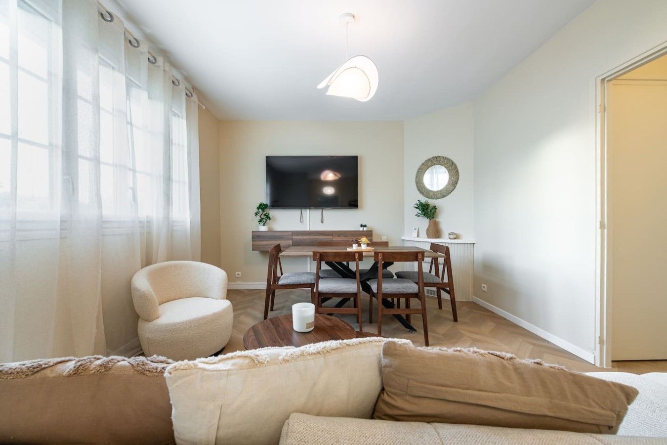Een woonkamer met een bank, een salontafel, een eethoek met tafel en stoelen, een wandgemonteerde flatscreen-tv en een deur aan de rechterkant.