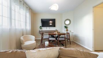 Een woonkamer met een bank, een salontafel, een eethoek met tafel en stoelen, een wandgemonteerde flatscreen-tv en een deur aan de rechterkant.
