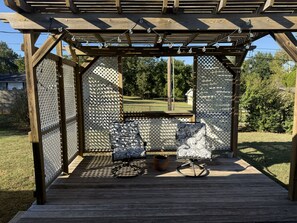 A wooden deck with two patterned chairs and a potted plant, under a pergola with string lights. - Your Home Base for OSU Game Days & Stillwater Stays (Stillwater)