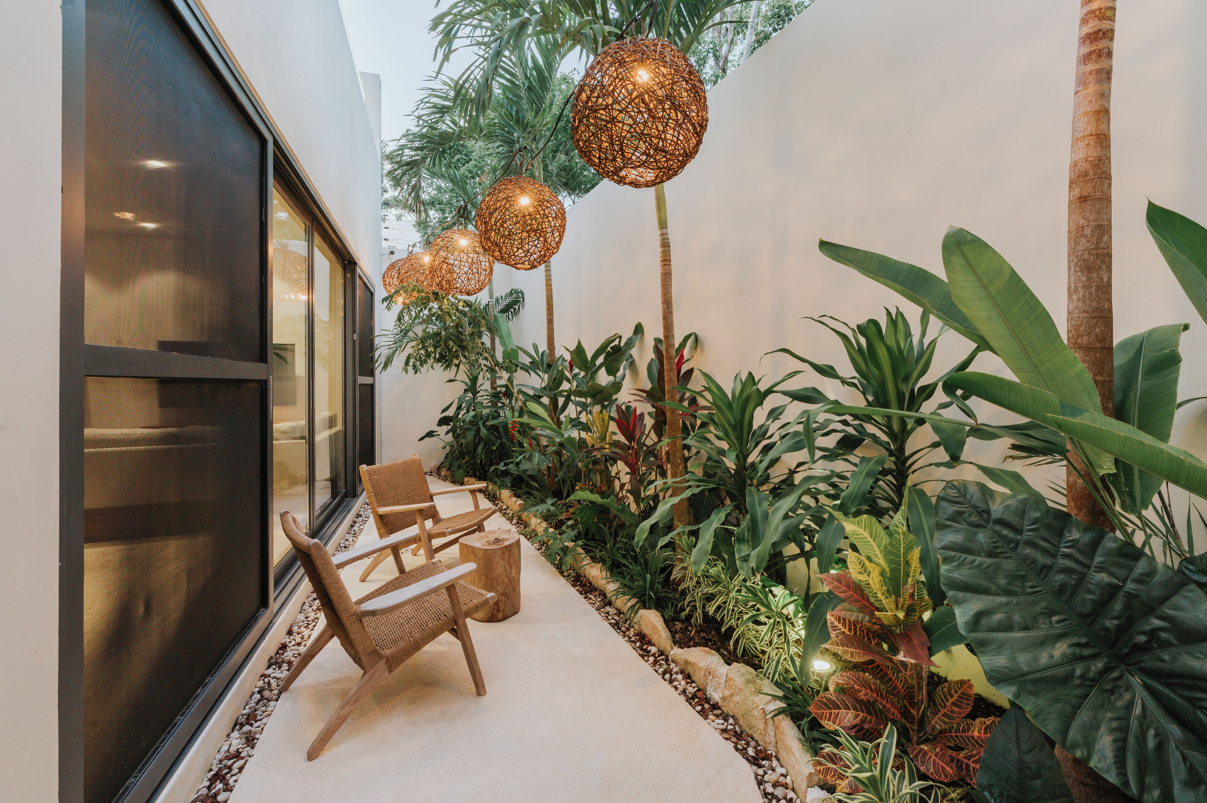 Uma área de estar moderna ao ar livre, com cadeiras de vime, parede de vidro e plantas tropicais.