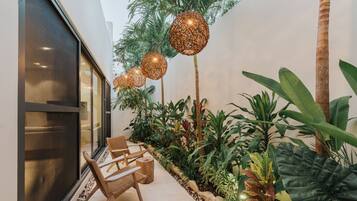Uma área de estar moderna ao ar livre, com cadeiras de vime, parede de vidro e plantas tropicais.