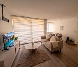 Ein modernes Wohnzimmer mit Fernseher, einem beigefarbenen Sofa, einem Couchtisch und einem großen Fenster mit Jalousien.