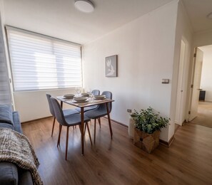 Ein Essbereich mit einem Tisch für vier Personen, einem Fenster mit Jalousien, einer wandmontierten Leuchte und einer Zimmerpflanze.