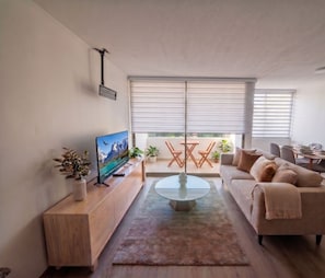 Ein modernes Wohnzimmer mit einem Flachbildfernseher, einem Glastisch, einem beigefarbenen Sofa und einem Essbereich mit Tisch und Stühlen.