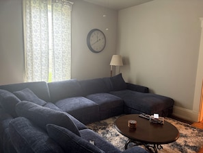 Una sala de estar con un sofá seccional, una mesa de centro redonda, un reloj de pared y un ventanal con cortinas estampadas.