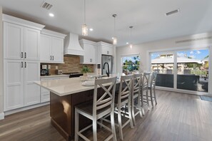 A modern kitchen with white cabinetry, a central island with bar stools, and a view of a beach house through a large sliding glass door. - 🌴Oasis on Ohio Ave | Canal-Front | Private Heated Pool + Steps to Beach 🌴 (Fort Myers Beach)