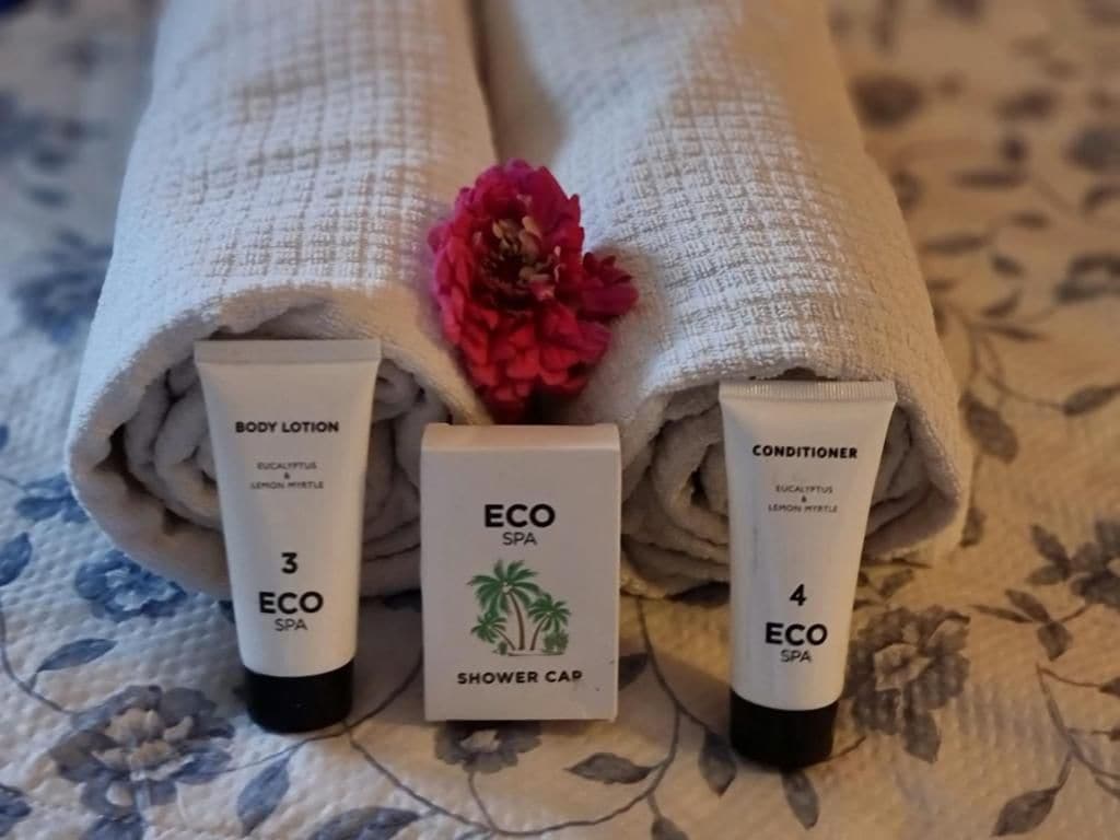 Trois serviettes roulées ornées d’un motif floral, une fleur rose et deux tubes de lotion corporelle et d’après-shampoing.