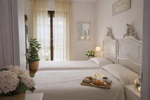 A hotel room with two beds, a bedside table with a lamp, a window with curtains, and a tray with breakfast items.