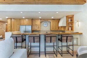 A modern kitchen with wooden cabinets, a central island with bar stools, and a clock on the wall. - Village Center 3G | Renovated 2BR in the Heart of Vail Village (Vail)